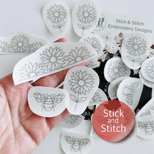 May include: A selection of white, pre-printed embroidery designs featuring sunflowers and bees. The designs are outlined in black ink and include the text "Stick & Stitch Embroidery Designs". The designs are held in a hand.