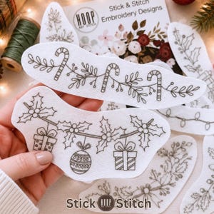 Könnte beinhalten: Weiße, selbstklebende Stickvliesbögen mit schwarzen Linienzeichnungen von weihnachtlichen Zuckerstangen, Stechpalmenblättern, Beeren, Geschenken und Christbaumkugeln. Die Verpackung zeigt den Text Hoop Art Stitch Stick & Stitch Embroidery Designs.