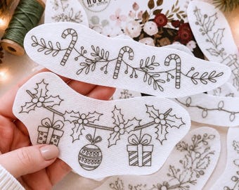 May include: White adhesive embroidery stabilizer sheets featuring black line art designs of Christmas candy canes, holly leaves, berries, gift boxes, and ornaments. The packaging displays the text Hoop Art Stitch Stick & Stitch Embroidery Designs.