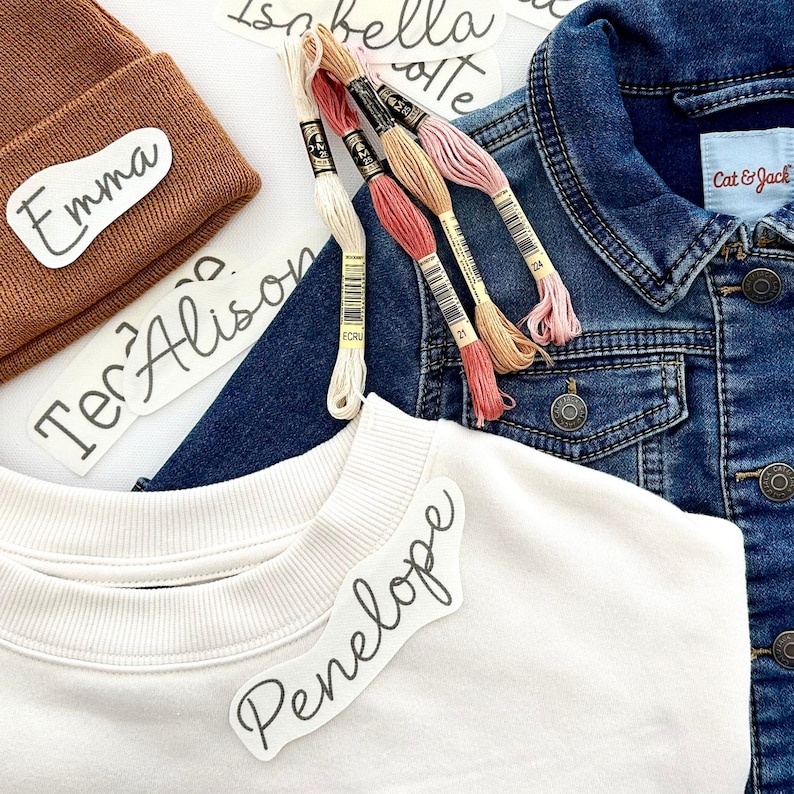 May include: A white sweatshirt with a black cursive name tag that says "Penelope" on it. There is a brown beanie with a white name tag that says "Emma" on it. There is a denim jacket with a white name tag that says "Cat & Jack" on it. There are also several spools of embroidery floss in various colors.