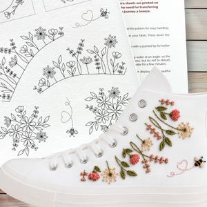 Puede incluir: Una zapatilla de lona blanca de ca&ntilde;a alta con un intrincado bordado floral en hilo rosa, amarillo y verde, junto a un peque&ntilde;o motivo de abeja y coraz&oacute;n. Junto al zapato hay una hoja de instrucciones con patrones florales en l&iacute;nea negra.