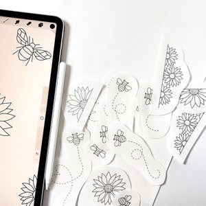 May include: A digital tablet screen displaying a black and white illustration of a bee and a sunflower. The tablet is on a white surface with several cut-out paper designs of bees and sunflowers.