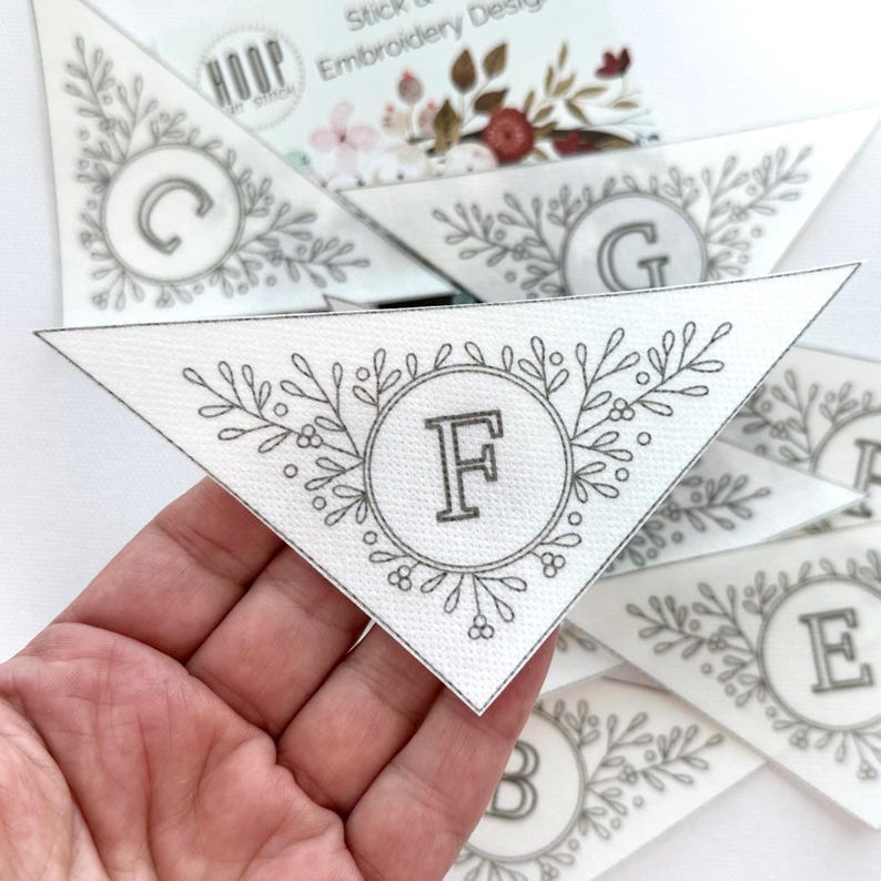 Puede incluir: Dise&ntilde;o de bordado triangular blanco con la letra "F" en un c&iacute;rculo gris, rodeado por una corona floral. Se ven otros dise&ntilde;os con letras. El dise&ntilde;o es para bordar.