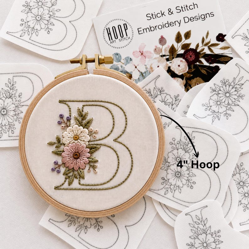 May include: A 4-inch wooden embroidery hoop containing fabric with a stitched letter B decorated with pink, white, and green floral embroidery. Surrounding the hoop are several white adhesive stick and stitch embroidery design patches.