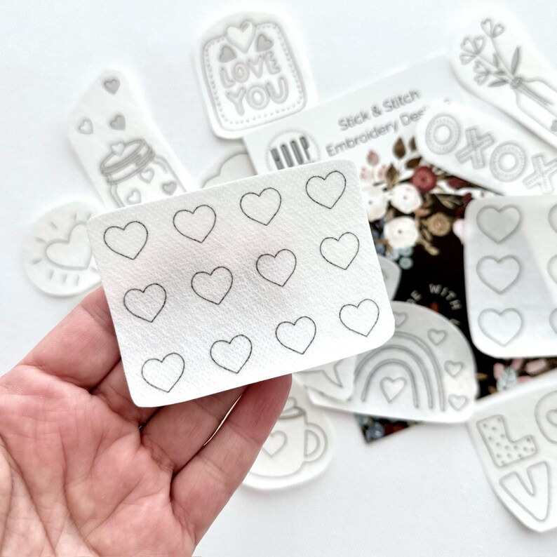 May include: A sheet of white paper with 16 outlined hearts for embroidery. The sheet is held in a hand. The sheet is part of a set of embroidery designs.