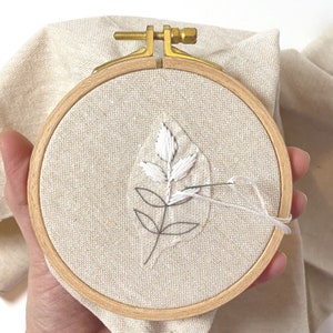 May include: A close-up of a white embroidery of a leaf on a beige fabric stretched in a wooden embroidery hoop.