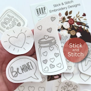 May include: A set of white iron-on embroidery designs featuring various heart-themed designs, including a jar filled with hearts, a heart with rays, a speech bubble with the text "Be MINE!" and a heart with the text "YOU LOVE". The designs are part of a "Stick & Stitch" embroidery collection.