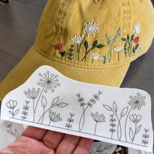 May include: A mustard yellow baseball cap featuring embroidered wildflowers in white, green, and red thread. Below the cap, a white paper template displays a black line drawing of the same floral pattern, showing the design layout for embroidery.