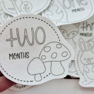 May include: A white felt baby milestone card with a black outline of a mushroom and the text "TWO MONTHS".