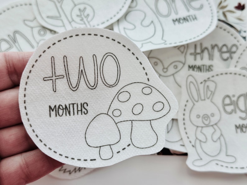 May include: A white felt baby milestone card with a black outline of a mushroom and the text "TWO MONTHS".