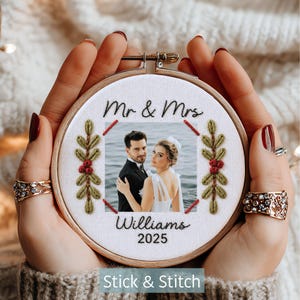 Könnte beinhalten: Ein runder hölzerner Stickrahmen mit weißem Stoff und einem zentralen Foto eines Paares. Der Text Mr & Mrs Williams 2025 ist in Schwarz gestickt. Zwei vertikale Zweige mit grünen Blättern und roten Beeren flankieren das Foto, das mit roten Stichen umrandet ist.