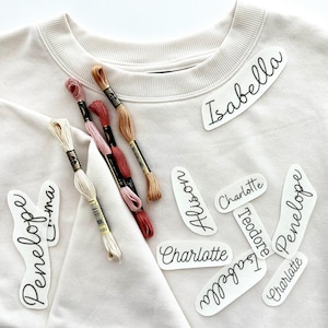 May include: A white sweatshirt with several white iron-on labels with names written in black cursive. The names include Isabella, Alison, Charlotte, Theodore, Penelope, and Emma. There are also several spools of embroidery floss in various colors.