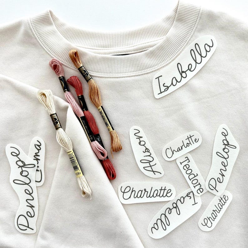 May include: A white sweatshirt with several white iron-on labels with names written in black cursive. The names include Isabella, Alison, Charlotte, Theodore, Penelope, and Emma. There are also several spools of embroidery floss in various colors.