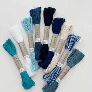 May include: A collection of Sashiko embroidery thread in various shades of blue, white, and cream. The spools are labeled with Japanese text and are arranged on a white surface. The threads are made of 100% cotton.