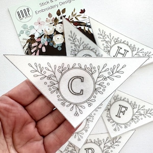 Peut inclure: Un ensemble de fanions triangulaires en tissu blanc avec des motifs de broderie noire. Chaque fanion pr&eacute;sente une lettre de l'alphabet dans une couronne de feuilles. Les lettres sont C, F et H.