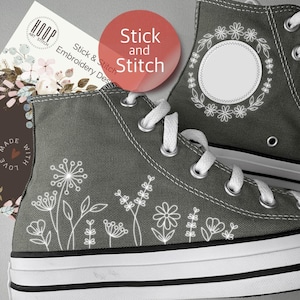 May include: Gray high-top sneakers with white floral embroidery. The sneakers have white laces and a white rubber sole with a black stripe. The text "Stick and Stitch" is on a red circle. A card with the text "HOOP ART STITCH" is visible.