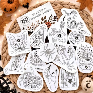 May include: A collection of white stick and stitch embroidery design patches featuring black line art illustrations of a cat, snake, spider, pumpkin, mushrooms, a cauldron, a crystal ball, a candle, and a hand, with an instruction card.