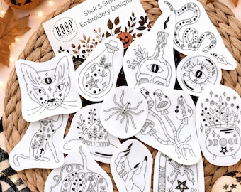May include: A collection of white stick and stitch embroidery design patches featuring black line art illustrations of a cat, snake, spider, pumpkin, mushrooms, a cauldron, a crystal ball, a candle, and a hand, with an instruction card.