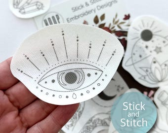May include: A white fabric sticker with a black and gray eye design. The eye has a crescent moon shape inside of it. The sticker is part of a set of embroidery designs called "Stick & Stitch Embroidery Designs".