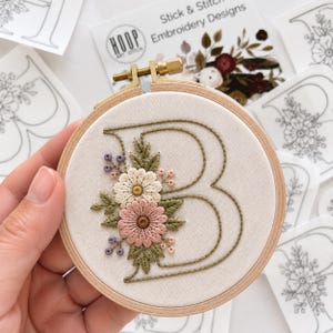 May include: A wooden embroidery hoop containing a cream-colored fabric with a hand-embroidered letter B in green thread, decorated with pink and white daisy-like flowers, green leaves, and small purple and peach-colored accent buds.