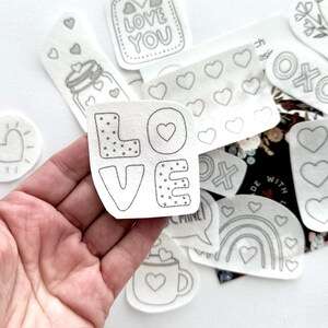 May include: A set of iron-on patches featuring various designs, including hearts, a rainbow, a mug, a speech bubble, and the word "LOVE" in a stylized font. The patches are white with black outlines and are designed for crafting and personalization.