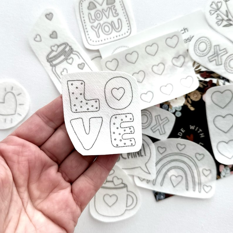 May include: A set of iron-on patches featuring various designs, including hearts, a rainbow, a mug, a speech bubble, and the word "LOVE" in a stylized font. The patches are white with black outlines and are designed for crafting and personalization.