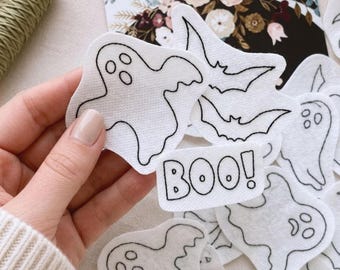 Könnte beinhalten: Eine Sammlung weißer Filzformen mit schwarzen Stickkonturen von Geistern, Fledermäusen und einem rechteckigen Aufnäher mit dem Text BOO! in fetter Schrift. Die Formen sind einzeln ausgeschnitten und weisen detaillierte Nähte an den Rändern auf.