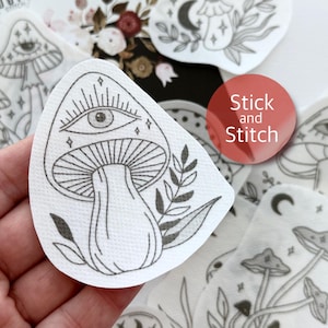 May include: A white sticker with a black line drawing of a mushroom with an eye on the cap. The mushroom is surrounded by leaves and stars. The sticker says "Stick and Stitch" in red text.