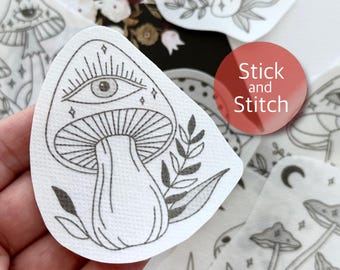 May include: A white sticker with a black line drawing of a mushroom with an eye on the cap. The mushroom is surrounded by leaves and stars. The sticker says "Stick and Stitch" in red text.