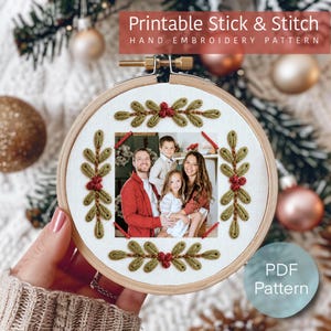 Peut inclure: Un tambour à broder en bois contenant un tissu blanc avec une photographie carrée d'une famille au centre. Le tissu est décoré de feuilles vertes et de baies rouges brodées en bordure rectangulaire. Le texte indique Printable Stick & Stitch.