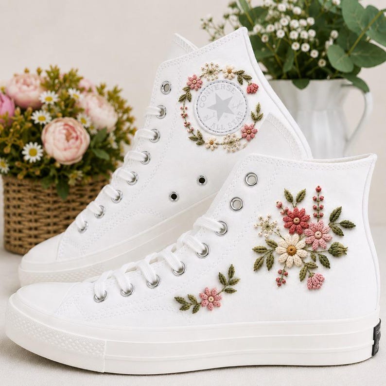 以下が含まれることがあります： 刺繍の花柄が施された白いハイカットスニーカーのペア。上のスニーカーには花輪で囲まれた円形のパッチがあり、下のスニーカーにはピンクとクリーム色の花と緑の葉のクラスターがデザインされています。