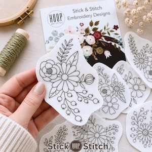 May include: A set of white adhesive embroidery patterns featuring black line art of floral bouquets with leaves and berries. The packaging is labeled Stick & Stitch Embroidery Designs by Hoop Art Stitch. These peel-and-stick sheets are for fabric.
