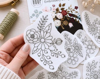 Op de afbeelding: Een set witte zelfklevende borduurpatronen met zwarte lijntekeningen van bloemenboeketten met bladeren en bessen. De verpakking is gelabeld met Stick & Stitch Embroidery Designs van Hoop Art Stitch voor gebruik op textiel.