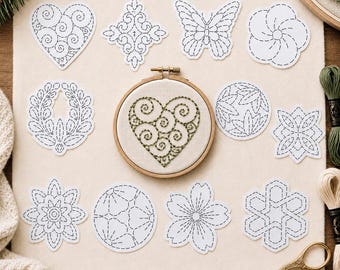 May include: A collection of twelve white fabric patches featuring blue dashed-line embroidery patterns, including hearts, flowers, butterflies, and geometric shapes, with one heart design already stitched in green thread inside a wooden embroidery hoop.