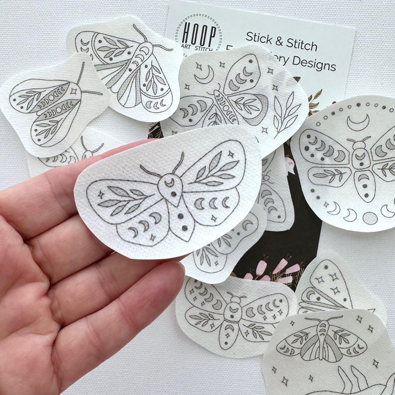 May include: A set of eight black and white moth embroidery patterns on white paper. The patterns feature moths with wings, antennae, and bodies, as well as moons, stars, and other celestial elements. The patterns are designed for stick and stitch embroidery.