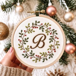 May include: A circular wooden embroidery hoop containing white fabric with a hand-stitched wreath design. The wreath features green leaves and small red berries encircling a large, brown, cursive letter B centered in the middle of the fabric.