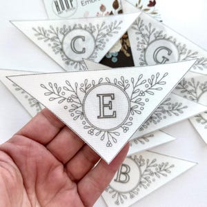 Puede incluir: Piezas triangulares de tela blanca con letras bordadas y dise&ntilde;os florales. La letra "E" es visible en un c&iacute;rculo, rodeada de hojas y bayas. Tambi&eacute;n est&aacute;n presentes otras letras, como "C", "G" y "B". Las piezas son parte de un kit de bordado DIY.