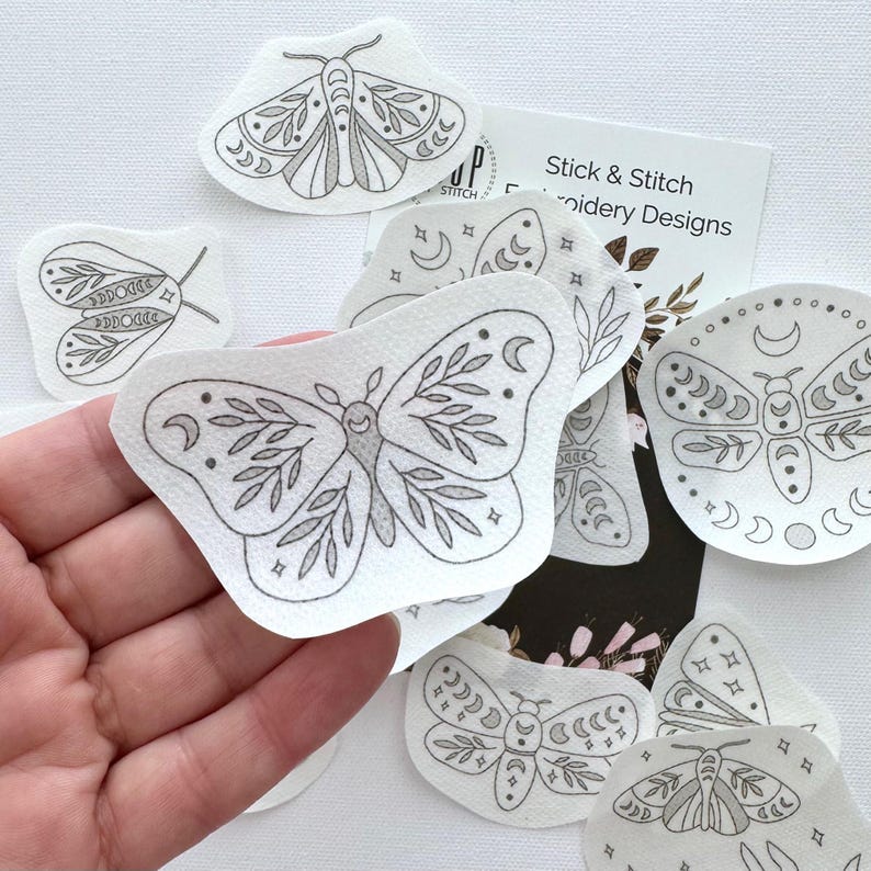 May include: A set of iron-on embroidery designs featuring moths and butterflies with moon and star details. The designs are printed on white fabric and are ready to be ironed onto fabric.
