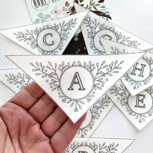 Peut inclure: Un ensemble de cinq fanions triangulaires en tissu blanc avec des broderies noires. Chaque fanion pr&eacute;sente une couronne florale avec une lettre au centre. Les lettres &eacute;pellent le mot "CACHE".
