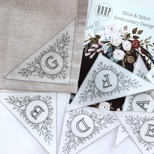 Peut inclure: Un ensemble de six pi&egrave;ces de tissu en forme de triangle avec un motif floral et les lettres A &agrave; G brod&eacute;es au centre de chaque pi&egrave;ce. Les pi&egrave;ces de tissu sont destin&eacute;es &agrave; un projet de broderie stick and stitch. Le texte "Stick & Stitch Embroidery Design" est visible sur une carte en arri&egrave;re-plan.
