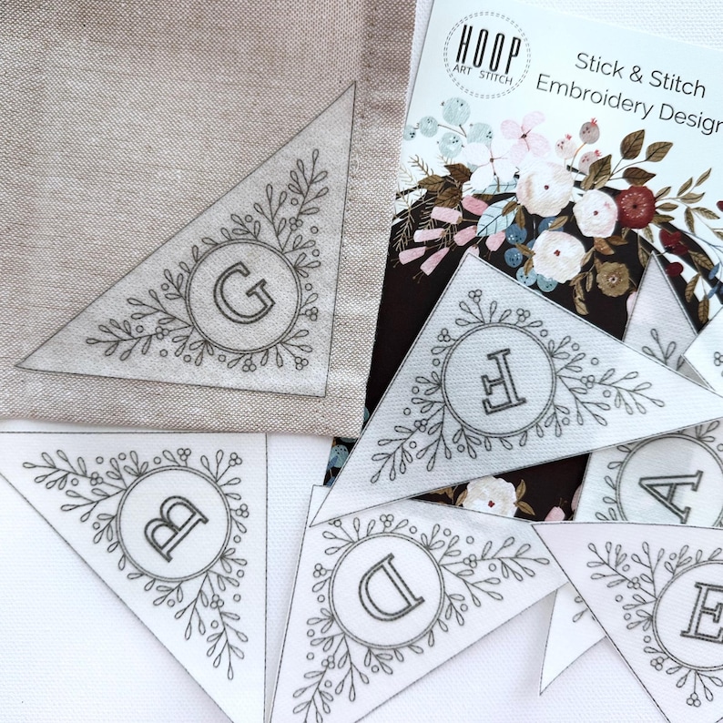 Peut inclure: Un ensemble de six pi&egrave;ces de tissu en forme de triangle avec un motif floral et les lettres A &agrave; G brod&eacute;es au centre de chaque pi&egrave;ce. Les pi&egrave;ces de tissu sont destin&eacute;es &agrave; un projet de broderie stick and stitch. Le texte "Stick & Stitch Embroidery Design" est visible sur une carte en arri&egrave;re-plan.