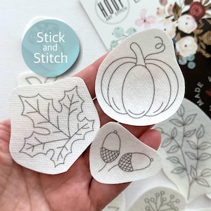 May include: Five white fabric patches with black outlines of a pumpkin, leaf, two acorns, and a sprig of leaves. The patches are labeled "Stick and Stitch".