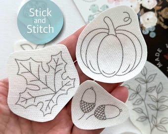 May include: Five white fabric patches with black outlines of a pumpkin, leaf, two acorns, and a sprig of leaves. The patches are labeled "Stick and Stitch".