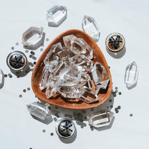 May include: A wooden bowl filled with clear quartz crystals. The bowl is surrounded by smaller crystals and decorative metal charms.