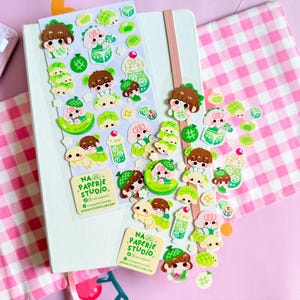 May include: A sheet of stickers featuring cartoon characters in green and brown tones, including small figures, melon-shaped items, and dice. The text NA PAPERIE STUDIO is printed on a square label at the bottom of the sticker sheet and on a separate tag.