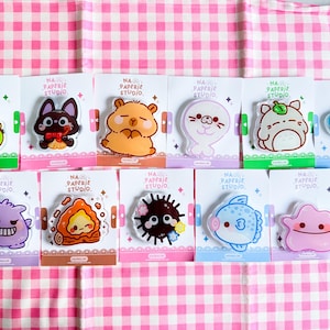 Può includere: Una collezione di undici spille in acrilico raffiguranti vari personaggi dei cartoni animati, tra cui una rana, un gatto, un criceto, una foca e creature marine. Ogni spilla è fissata su un cartoncino bianco con l'etichetta NA PAPERIE STUDIO.