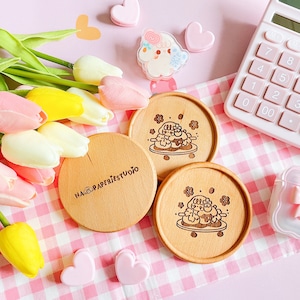 May include: Three round wooden coasters with a laser-etched design of a cartoon character on a plate. One coaster has the text "NA PAPERIE STUDIO". The coasters are on a pink and white checkered surface, with tulips, a calculator, and other accessories.