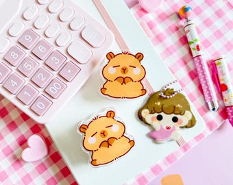 May include: A collection of cute stationery items, including three hamster-shaped stickers, a girl-shaped charm, and two pens with pink accents. A pink calculator and a heart-shaped eraser are also visible, all arranged on a pink and white checkered surface.
