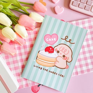 Può includere: Un quaderno verde pastello a righe verticali con un'illustrazione di un cane che tiene una torta. Il testo "Cake" è sopra la torta e "Living the sweet life!" sotto. Tulipani rosa.