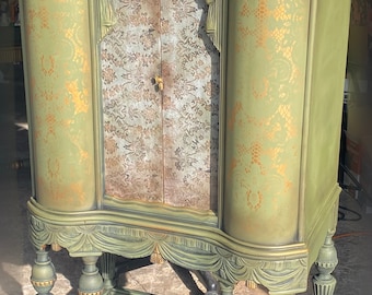 May include: A vintage green cabinet with gold accents and a decorative lace pattern. The cabinet has a curved top and ornate legs.